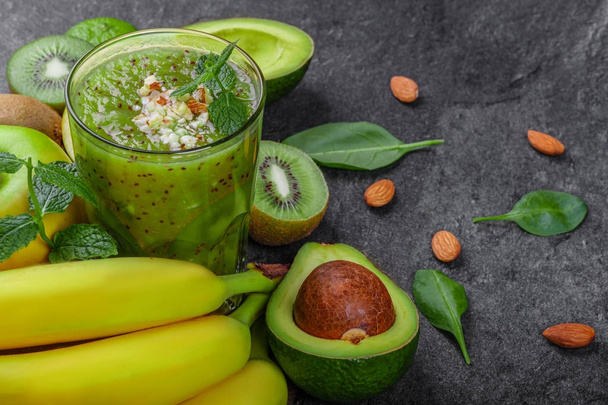 Close-up of different exotic fruits on a gray stone background. A heap of bananas, nutritious avocados, juicy kiwis and peppermint. A tall glass of kiwi beverage with spices and nuts. - Photo, image