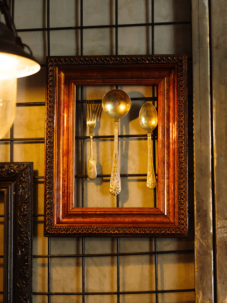 Shiny decorative spoons and silver fork in a brown wooden frame hanging on a cafe wall background. A set of antique utensil for bar or pub decoration. Luxurious ethnic spoons and forks. - Foto, immagini