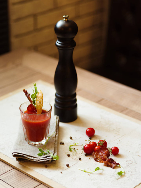 A top view of a glass of Bloody Mary and cherry tomatoes on a white fabric and on a wooden table. Stylish pepper mill and alcohol cocktail with bacon on a top of glass on a blurred background. - Valokuva, kuva