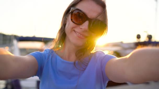 Жінка в сонцезахисних окулярах робить селфі на тлі пристані з великою кількістю яхт і човнів на заході сонця, крупним планом. Повільний рух
 - Кадри, відео