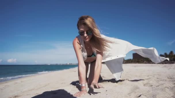 Chica, playa, mar, viento en tu cabello Una hermosa chica en bikini de color y una capa blanca transparente, se sienta en la arena sobre sus rodillas y levanta sus manos con arena fina de la playa y la vierte
 - Metraje, vídeo