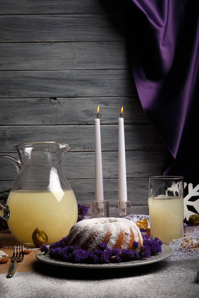A beautiful table setting from holidays food on a gray wooden background. Traditional homemade cake with sweet icing and decorative flowers on a plate. Pineapple juice and long candles on a table. - Photo, image