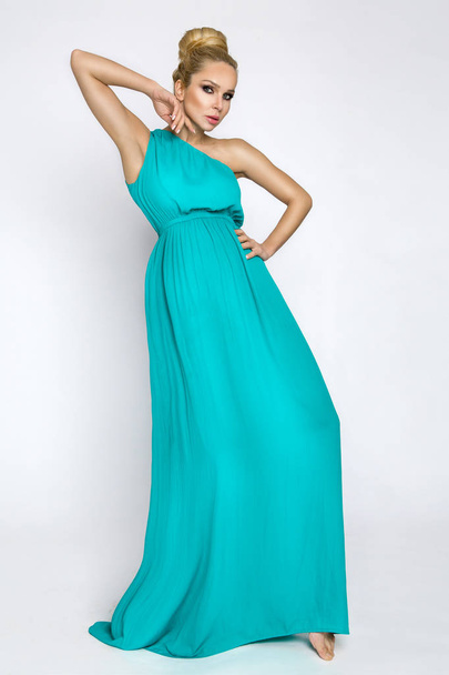 Elegante vrouwelijke model in een turquoise lange zijden jurk staande op een witte achtergrond en sensueel vormt - Foto, afbeelding