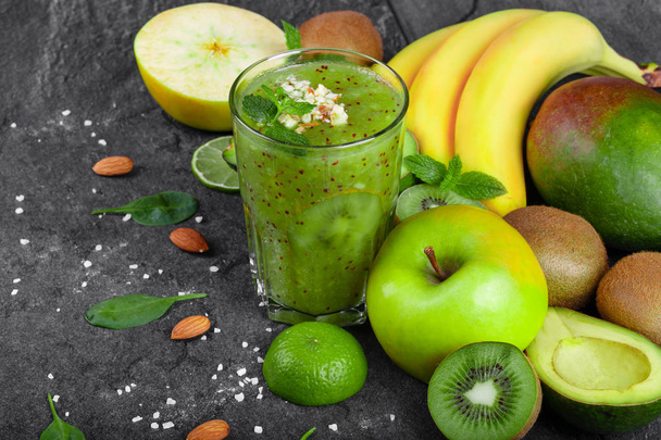 A top view of a colorful composition of a kiwi smoothie with grated almond and tropical fruits on a dark stones background. Whole and cut apples, kiwi, avocado and a branch of bananas on a table. - 写真・画像