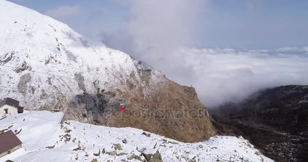 εναέρια πετώντας πάνω από το χιονισμένο βουνό κορυφογραμμή κοιλάδων με σύννεφα, κόκκινο καλώδιο αυτοκίνητο ανύψωση Θεοδωρούδη. Μεταφορά γόνδολα τουριστών αλπική άγρια φύση εξωτερική χιόνι το χειμώνα. 4 k κηφήνας πτήση πλάνο για τη θέσπιση - Πλάνα, βίντεο