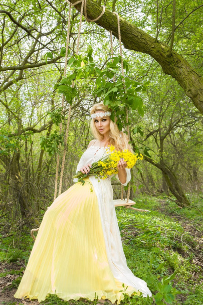 Потрясающая блондинка сексуальная натуральная женщина качается на качелях на дереве в весеннем платье, держа букет весны
 - Фото, изображение