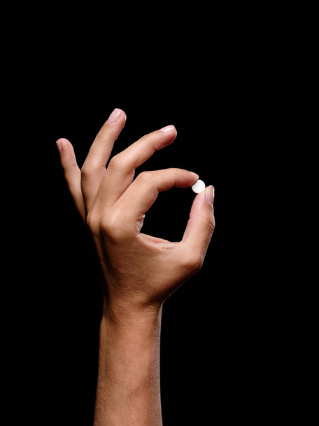 A man's hand holding a little round white pill between thumb and forefinger on a black background. Prescripted medication: painkillers, light narcotics, antibiotics. Pharmaceutical modern science. - Foto, afbeelding