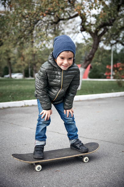 Cute funny smiling little boy wearing dark grey jacket, dark blue cap and blue jeans, riding on grey skate board in garden  - Photo, image