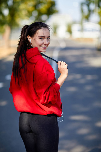 A sporty young woman, in a red sweatshirt with headphones in her ears and a skipping rope in her hands. - Φωτογραφία, εικόνα
