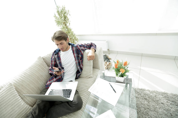 ノート パソコンとスマート フォンのリビング ルームのソファの上に座っている若い男. - 写真・画像