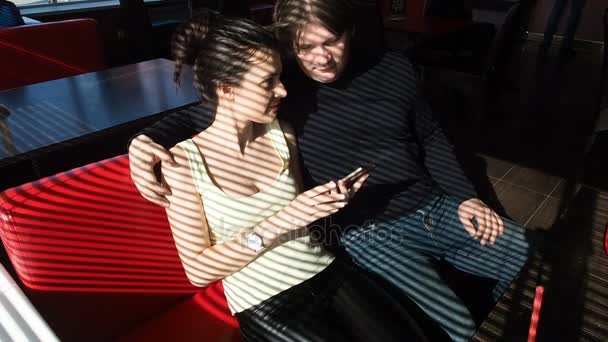 Woman and man are sitting at table in cafe. Girl showing something on mobile phone and smiling. The guy hugs her shoulders. Cute conversations in a cafe - Footage, Video