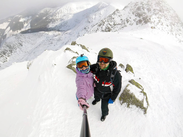 молодой и счастливый парень и девушка делают фото с палкой и смартфоном на вершине заснеженной горы на горнолыжном курорте
 - Фото, изображение