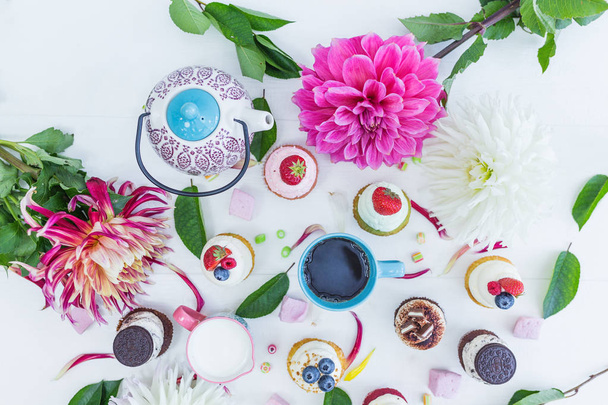 Various cupcakes with fresh berries flowers and leaves, a cup of tea or coffee and a kettle. Top view. - Photo, Image