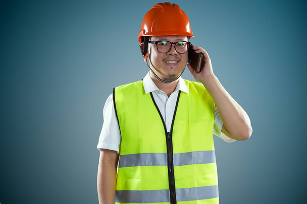 Young asian construction worker in safety helmet and using smartphone, isolated on blue background - Foto, imagen