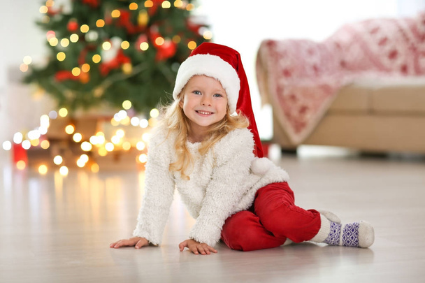 Cute little girl in Christmas hat sitting on floor at home - 写真・画像