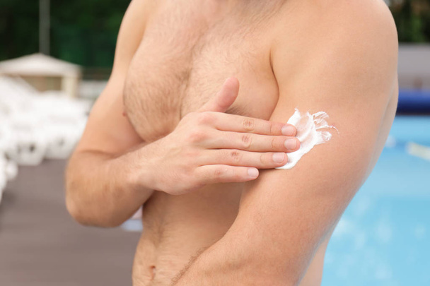 Komea nuori mies soveltamalla aurinkovoidetta iholle lähellä uima-allas
 - Valokuva, kuva
