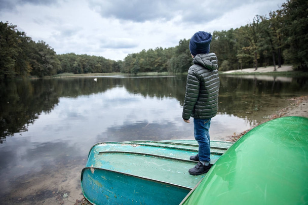 Little cute boy wearing dark jacket, blue jeans, black shoes and dark blue hat, standing on the boat on the bank of the lake  - Fotó, kép