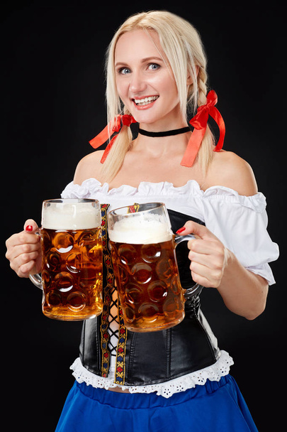 Jonge sexy vrouw draagt een dirndl met twee bier mokken op zwarte achtergrond. - Foto, afbeelding