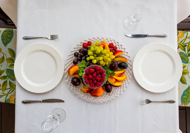 Тарелка фруктов с малиной, виноградом и сливой на столе на двоих
 - Фото, изображение
