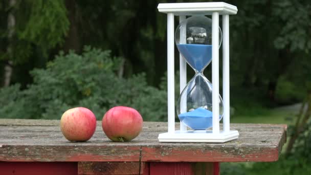 Bodegón de fin de verano con vidrio de arena de reloj de arena azul y dos manzanas en la mesa vieja en el jardín
 - Metraje, vídeo