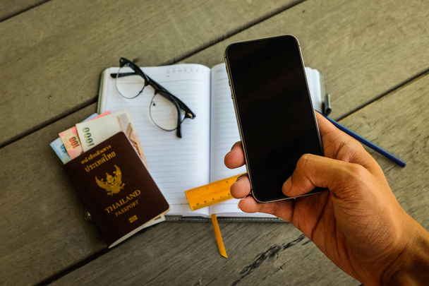 Mão de homem usando telefone celular (tela preta) com passaporte da Tailândia, dinheiro, régua amarela, lápis, notebook e óculos para negócios de contato e preparar viagens
. - Foto, Imagem