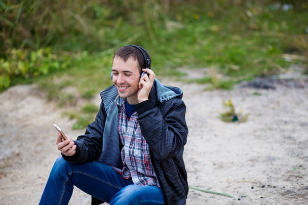 Genç adam ruhları nehir kıyısında bir telefon ile oturur ve müzik dinliyor - Fotoğraf, Görsel