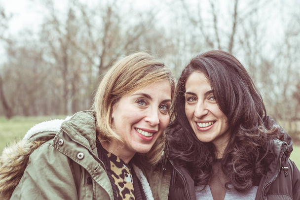 Dwa piękne młode kobiety mówią, uśmiechając się i zabawy na świeżym powietrzu w zimny, jesienny dzień. Dostęp do światła naturalnego, skóry naturalne, naturalne tło. - Zdjęcie, obraz