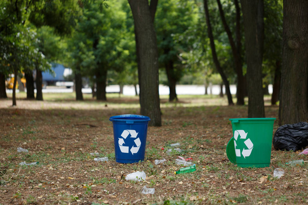 A couple of bright blue and green recycling bins in the park. Plastic rubbish containers next to plastic bottles on a blurred natural background. Environment, ecology, recycling, pollution concept. - Fotó, kép