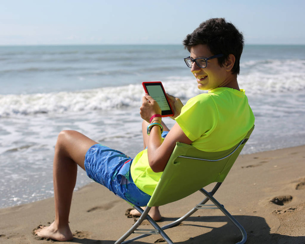 s úsměvem kavkazské chlapec čte ebook sedí na pláži židli - Fotografie, Obrázek