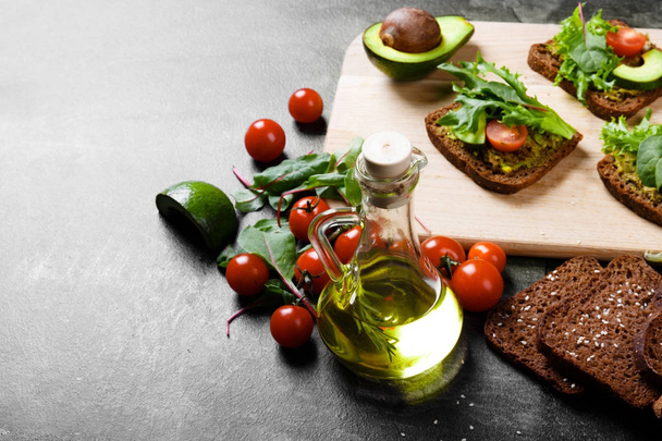 Close-up picture of a thick olive oil, cherry tomatoes, greens and wheat bread for a healthful sandwich. Toasted avocado sandwiches for breakfast on a cutting deck on a shiny table background. - Zdjęcie, obraz