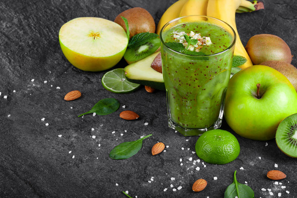 A colorful composition of an organic kiwi smoothie with grated almond and tropical fruits on a dark stones background. Whole and cut apples, kiwi, avocado and a branch of yellow bananas on a table. - Фото, изображение