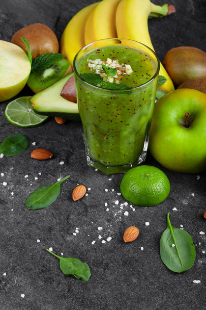 A top view of a composition of kiwi smoothie with grated nuts on a top and ripe fruits on a dark stones background. Whole and cut apples, lime, kiwi, avocado and a branch of ripe bananas on a table. - Valokuva, kuva