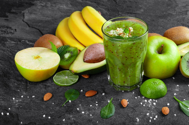 A colorful composition of kiwi smoothie with grated nuts on a top and ripe fruits on a dark stones background. Whole and cut apples, lime, kiwi, avocado and a branch of ripe bananas on a table. - Photo, image