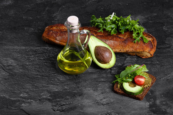A top view of a glass jar full of an avocado oil and freshly baked baguette on a dark gray stone background. Healthful sandwich with organic vegetables and a cut avocado on a table. Copy space. - Foto, Imagem