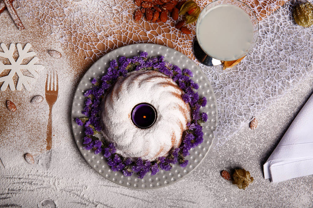 A top view of a composition of a traditional Christmas cake with icing and violet flowers around the baked dish on a plate and colorful background. A coconut beverage and a heap of almond on a desk. - Photo, image