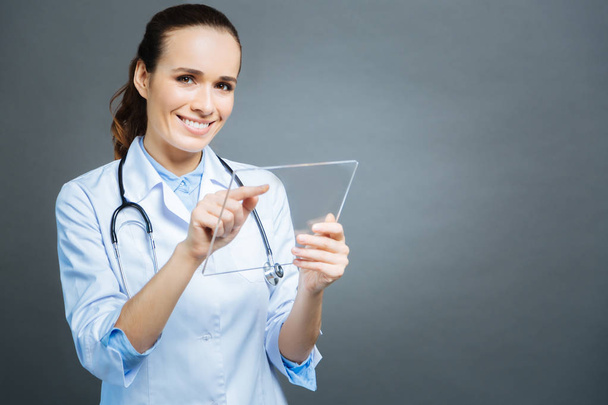 Beaming young lady in white coat posing with transparent tablet - 写真・画像