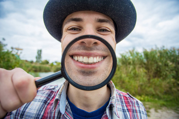 Een vrolijke man met een grappig gezicht in een hoed houdt een vergrootglas - Foto, afbeelding