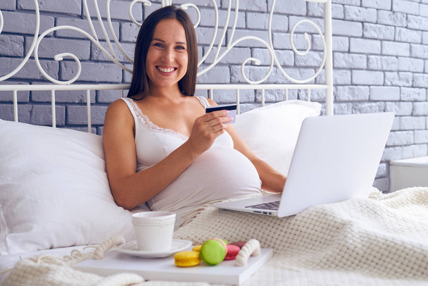Femme enceinte assise sur le lit avec ordinateur portable et carte de crédit
 - Photo, image