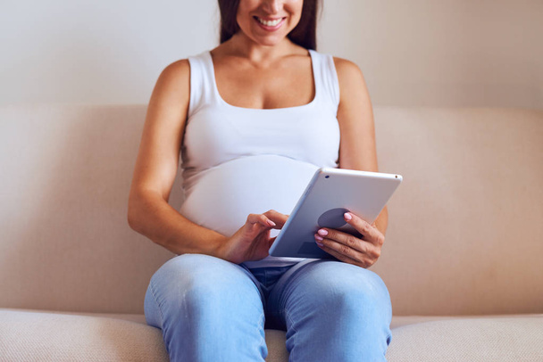 Ευτυχισμένη γυναίκα έγκυος χρησιμοποιώντας tablet ενώ κάθεται στον καναπέ  - Φωτογραφία, εικόνα