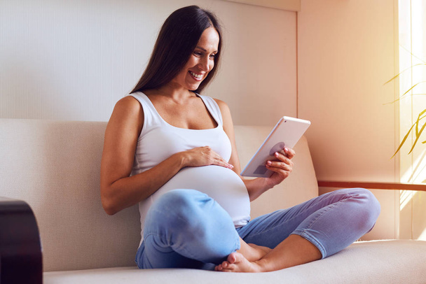 Νεαρή έγκυος γυναίκα που αναπαύεται με tablet στον καναπέ στο σπίτι - Φωτογραφία, εικόνα