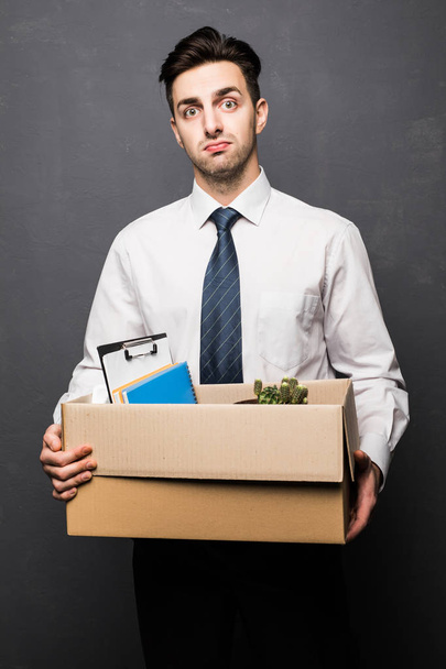 Getting fired. Handsome businessman in formal wear holding a box with his stuff, on gray background - Fotó, kép