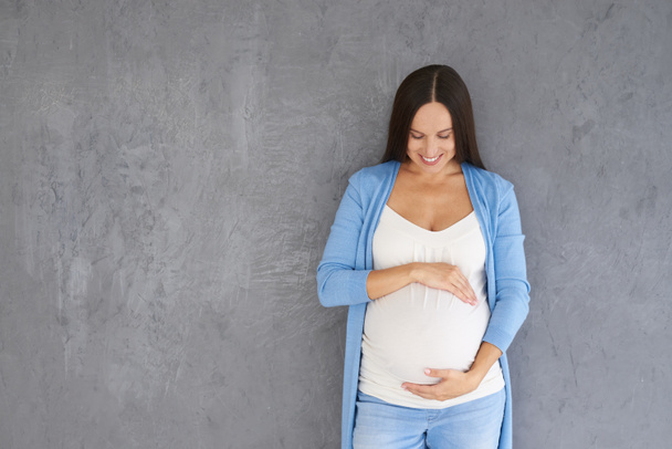 Молодая беременная женщина смотрит на свой живот против серой спинки
 - Фото, изображение