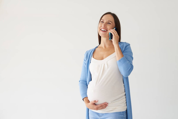 Smiling pregnant woman talking on smartphone against white backg - Фото, зображення