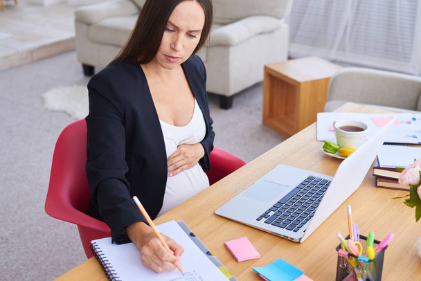 Empresaria embarazada enfocada cogida de la mano en el vientre durante el trabajo
 - Foto, imagen