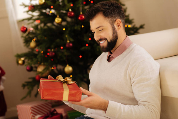 Pleasant man holding a gift box and smiling - Fotó, kép