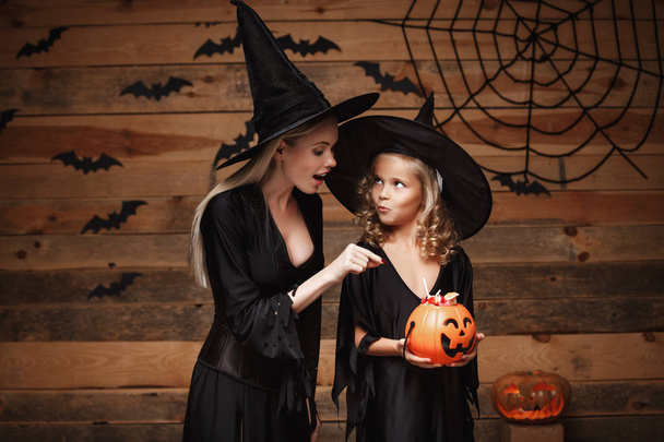 Concept d'Halloween - belle mère caucasienne et sa fille en costumes de sorcière célébrant Halloween en partageant des bonbons d'Halloween et doux sur chauves-souris et toile d'araignée sur fond de studio en bois
. - Photo, image