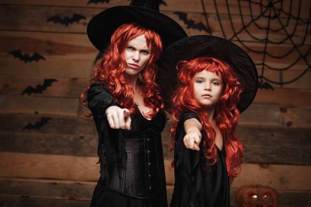 Cadılar Bayramı kavramı - güzel beyaz anne ve kızıyla uzun kızıl saçlı cadı kostümleri kızgın telaşlı yüze ilişkin tabir ve kamera için işaret parmağı ile birlikte. - Fotoğraf, Görsel