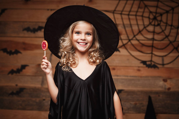 Halloween-Hexenkonzept - kleines Hexenkind mit Halloween-Süßigkeiten und Bonbons mit fröhlichem Lächeln. über Fledermaus und Spinnennetz Hintergrund. - Foto, Bild