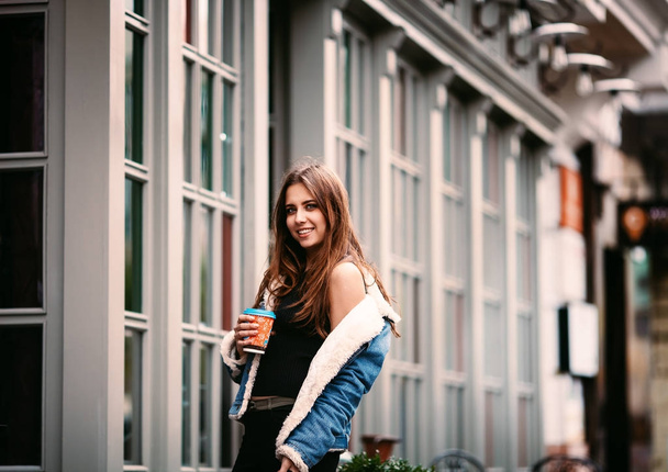 The concept of street fashion. portrait of Young girl dressed in a fashionable outfit. Posing against the window of the boutique girl smiling and drinking coffee - Photo, image