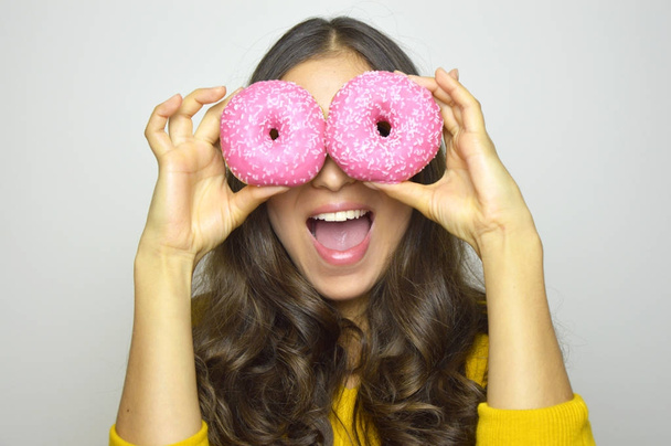 Close-up portrait of long-haired smiling girl having fun with sweets isolated on gray background. Attractive young woman with long hair posing with pink donuts in her hands - Foto, Imagen